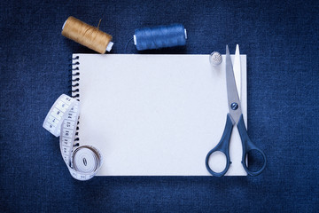 Scissors, Thread, Thimble, Centimeter Tape On Dark Blue Gray Jeans Fabric With Cardboard Sheet Of Notebook Copyspace Top View.