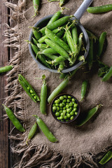 Young organic green pea pods and peas in can tin and aluminum colander over old dark wooden planks with sackcloth textile background. Top view with space. Harvest, healthy eating.
