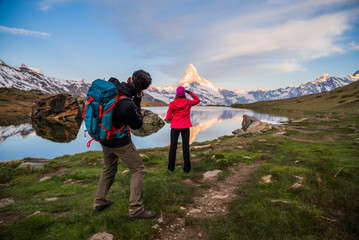 Excited young woman standing in front of a lake photographed by her boyfriend. Photographer taking portraits of his girlfriend admiring the Swiss Alps