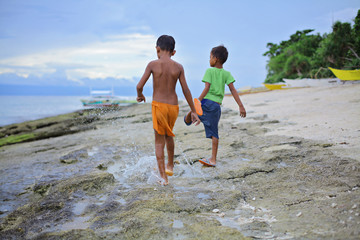 two little asian boys splashing their feet in shallow water ahead of oncoming waves of the sea at sunset, Island, Philippines