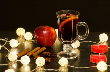Mug of mulled wine with spices, candles in the shape of a heart, cinnamon sticks, star anise. Illumination of rattan lanterns on a black wooden table