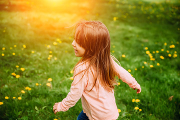 Little girl running along the green lawn with yellow dandelions, back view. Background image, concept on the theme of happiness, childhood and carefree.