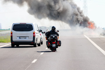 Car crash / accident / road disaster concept. Vehicles passing a car burning with a thick black...