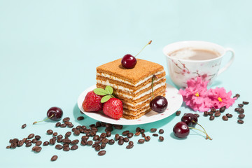 Tasty cake on the white plate and cup of coffee with cream, decorated with flowers, coffee beans, strawberries and ripe cherry on the light mint background with place for text, perspective view