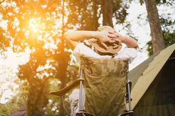  Back side of young man tourist sitting on chair resting and relaxing in front of tent at camping site in forest. Outdoor activity in summer. Adventure traveling in national park.