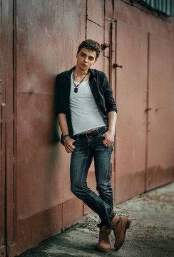 The young guy is standing near the garage. Poses for men's photo sessions. Beautiful appearance, clothes in rock style.