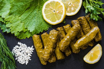 Delicious stuffed grape leaves (the traditional dolma of the mediterranean cuisine) on black dish with leaves, lemon slices, rice, parsley and dill - obrazy, fototapety, plakaty
