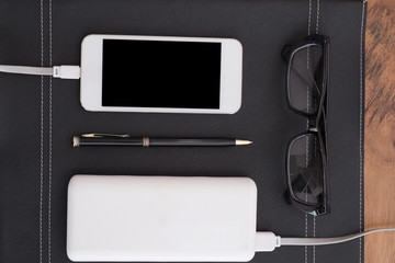 mobile phone charging from power bank, eyeglasses and pen on the black noetbook