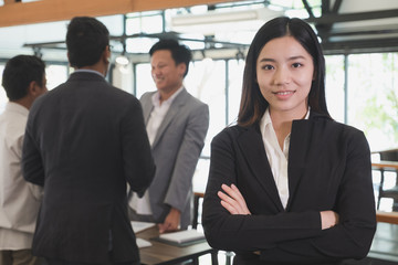 asian businesswoman with folded hands smiling at camera. confident young woman wearing black suit with crossed arms standing at office with businessman have a meeting behind her.