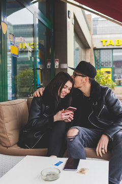 Young couple sitting in bar, he is kissing her head and she is using smart phone - tender, romance, happiness concept