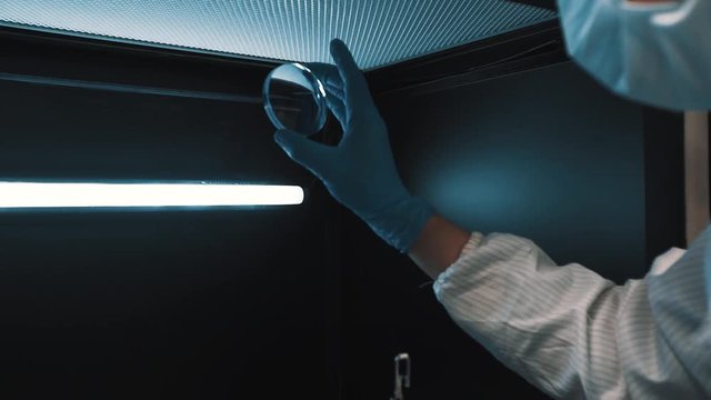 Scientist in sterile white robe blue rubber gloves manages lenses in metal lighted box at laboratory