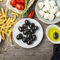 Healthy comfort food: ingredients for cooking pasta, salads, olives, sweet onions, mozzarella cheese, olive oil, juicy tomatoes on a plain wooden background. From the top view. The Mediterranean diet