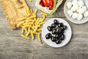 Healthy comfort food: ingredients for cooking pasta, salads, olives, sweet onions, mozzarella cheese, olive oil, juicy tomatoes on a plain wooden background. From the top view. The Mediterranean diet