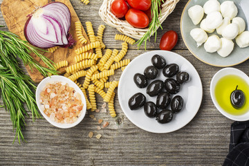 Healthy comfort food: ingredients for cooking pasta, salads, olives, sweet onions, mozzarella cheese, olive oil, juicy tomatoes on a plain wooden background. From the top view. The Mediterranean diet