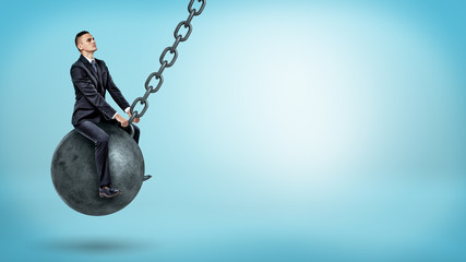 A businessman swinging on a large wrecking ball and looking up on blue background.