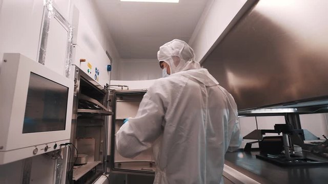 Engineer in sterile hazard clothes white robe and rubber gloves puts metal element inside manufacture oven in laboratory