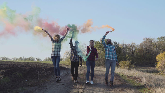 Four happy young multi racial friends in rural road under blue sky with colored smoke grenades.