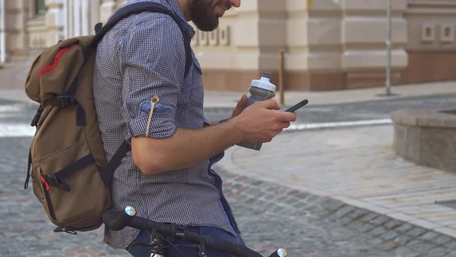 Attractive caucasian cyclist drinking water on his bicycle. Handsome brunette guy browsing internet on his smartphone. Bearded young man holding cellphone in one hand and bottle in another