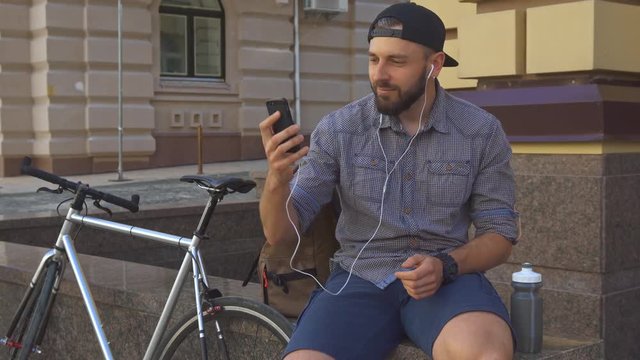 Handsome bearded cyclist having a video chat on his smartphone on the street of the city. Young caucasian man sitting on the edge near his bicycle. Attractive brunette guy in cap waving his hand for