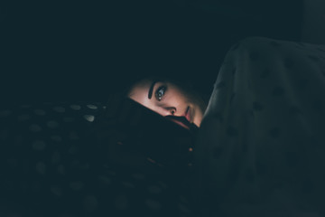 Woman using smart phone lying bed with face illuminated by screenlight - cheating, phubbing,...
