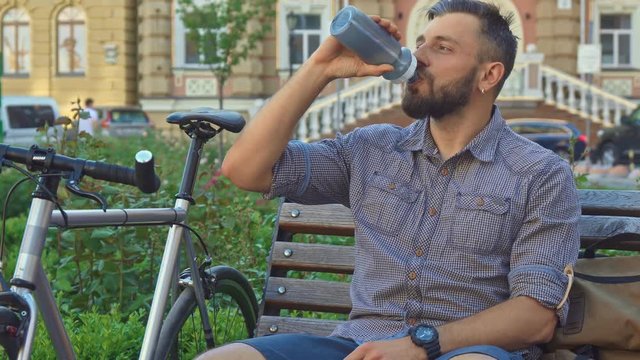 Young brunette cyclist drinking water on the bench. Handsome caucasian man taking a rest near his bike. Attractive caucasian guy looking to the sides against background of public building
