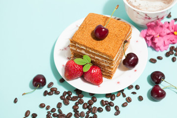 Tasty cake on the white plate and cup of coffee with cream, decorated with flowers, coffee beans, strawberries and ripe cherry on the mint background with place for text, top close up perspective view