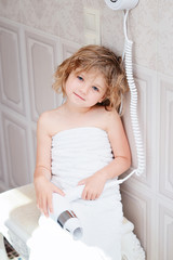 Obraz na płótnie Canvas Little girl in a towel sits in the bathroom with a hair dryer