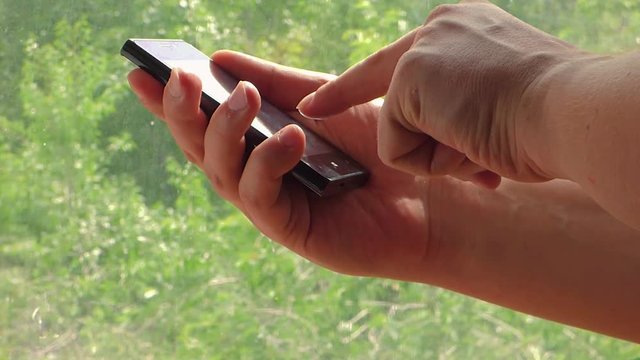 Closeup view of hands with black glossy smartphone. Caucasian man touch and scroll mobile phone screen. With blurred trees behind window at the back. Sunny day.