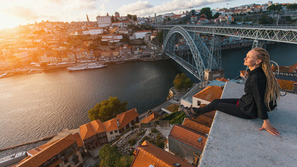 Young pretty woman with blond dreadlocks meets the sunset sitting on the view point in front of the Douro river and Dom luis I bridge in Porto, Portugal.