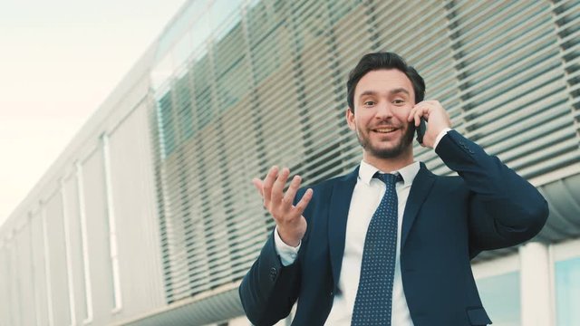 Hadsome businessman having a nice conversation on the cell phone. Hearing good news. Man looking at the camera with happy emotions. Outdoor shot. close up