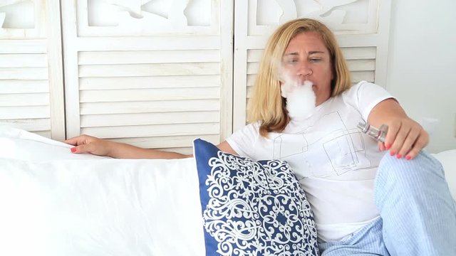 Portrait of attractive blonde woman laying on a bed smoking electronic cigarette at home interior