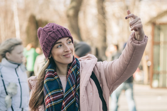 The attractive girl in a dark checkered scarf, a violet cap and a pink jacket walks in the winter. She lovely smiles and allegedly takes a selfie by means of a hand.