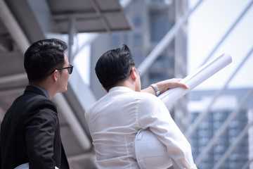 Asian businessman discuss with engineer architect in suite, looking and point to finger forward away and holding construction roll plan for working team concept,back view