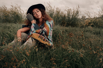 Beautiful boho woman wearing hat and poncho sitting on green field