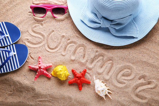 Word Summer written by hand on beach sand with hat, sunglasses and starfish