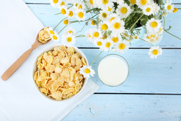 Cornflakes in bowl with glass of milk and chamomile on blue wooden table