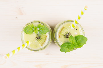 Freshly blended green kiwi fruit smoothie in glass jars with straw, mint leaf, top view. White wooden board background, copy space.