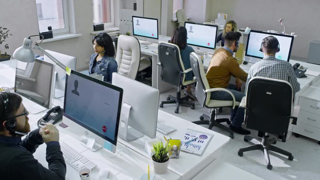 Crane shot of helpdesk operators in headsets working in office: working on computers and talking with customers on voice calls 