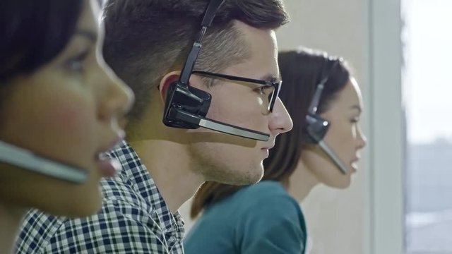 Medium shot of male call centre operator in glasses sitting beside female colleagues and talking to clients on headset, then PAN to reflection of female Asian colleague working 