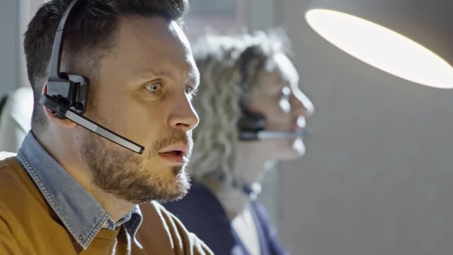  PAN of concentrated man with beard wearing headset with microphone and talking on voice call with client in call centre office; colleagues working beside him 