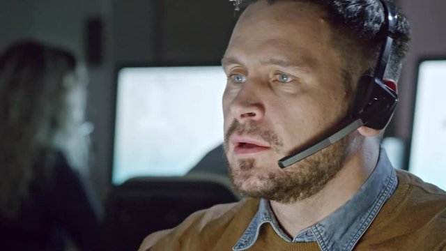 PAN of bearded man sitting before computer in dark call centre office and talking on headset with client 