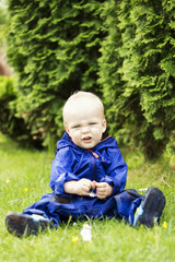 Funny baby boy sitting on the grass in a blue raincoat. Cute infant kid playing on the backyard