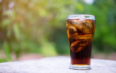 Glass of cola with ice cubes on table with green nature background. Copy space and texture.