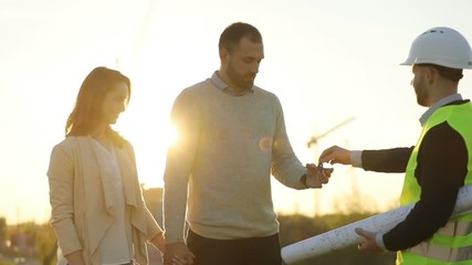 Side view of architect in white helmet giving keyses to young couple of clients, shaking hand of male client on sunset background. Outdoor.