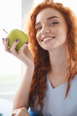 Cheerful redhead lady sitting at the table indoors eating apple