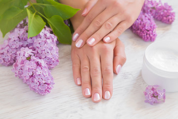 Fototapeta premium Hands moisturizing cream cares about woman's hands skin. Manicure beauty salon. Spring and summer gentle atmosphere with fresh and fragrant purple lilac flowers on the wooden table.