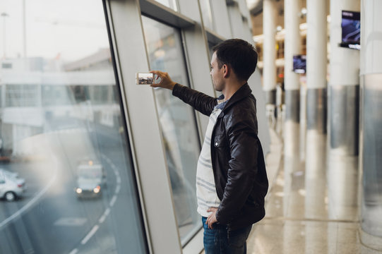 Handsome man standing beside glass wall in modern airport terminal, taking photo picture of airplane aircraft, travelling to other countires.