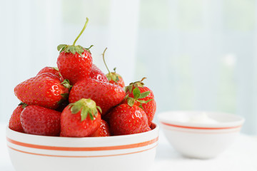 Many red ripe jucy strawberry at white plate close to cup of sour cream on table in light room