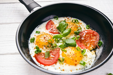 Pan of fried eggs with tomatoes, cheese, spring onion, herbs on a white table. White wooden table. Concept of food. Breakfast time. Copy space. 