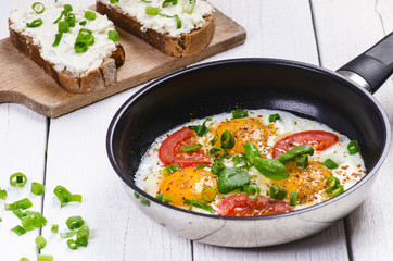 Pan of fried eggs with tomatoes, cheese, spring onion, herbs on a white table. Bread with spread. White wooden table. Concept of food. Breakfast time. Copy space. 